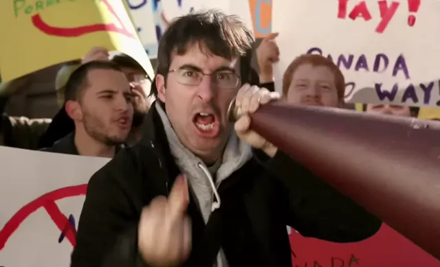 A man resembling John Oliver shouts intensely into a megaphone at what appears to be a protest, surrounded by people holding signs including one that reads 'Canada Way.' The scene is energetic and satirical in tone, likely from a comedy sketch.