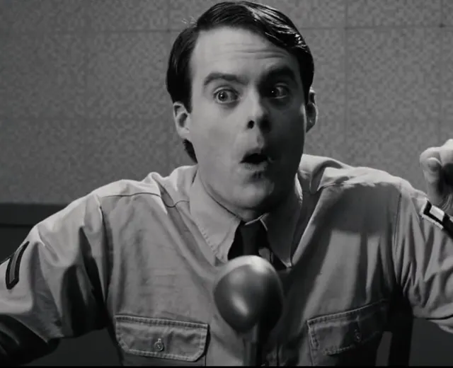 A man in a military-style uniform leans toward a microphone with a wide-eyed, open-mouthed expression of extreme shock or disbelief, captured in black and white. The scene appears to be from a film or TV production with a mid-20th century aesthetic.