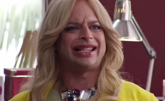 A person in drag with long blonde hair, heavy makeup, a yellow outfit, and a statement necklace displays an exaggerated crying or distressed facial expression. The setting appears to be an indoor scene, possibly from a TV show or comedy sketch.
