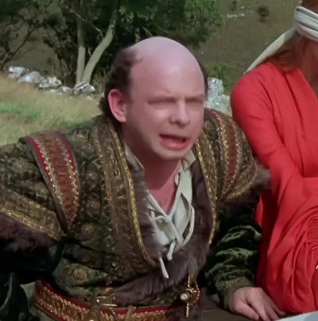 A bald man in ornate medieval-style robes appears to be speaking with an exasperated or scheming expression, recognizable as the character Vizzini from The Princess Bride. He is crouching or leaning forward outdoors.