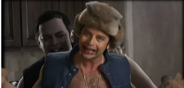 A vampire character bites or grabs a flamboyantly dressed man wearing a fur hat, gold chains, and a denim vest in what appears to be a comedic live-action or video game cutscene. The victim has an exaggerated, distressed expression.