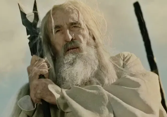 An elderly wizard with long white hair and beard, dressed in tattered white robes and clutching a dark staff, looks upward with a weary and determined expression. This appears to be Gandalf the White from the Lord of the Rings films during a battle sequence.