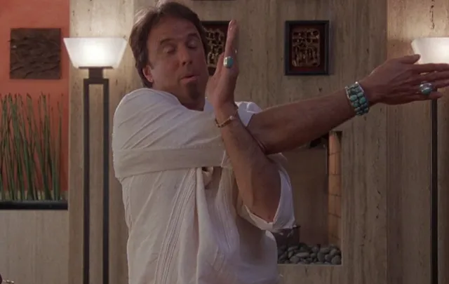 A middle-aged man in a white outfit adorned with turquoise jewelry strikes a dramatic pose with his arms outstretched and hands gesturing expressively, appearing to be in a mid-conversation or performative moment inside a modern interior setting.