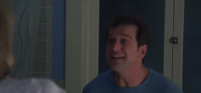 A middle-aged man in a blue t-shirt appears to be speaking animatedly or reacting with surprise to someone partially visible in the foreground. The setting looks like an interior hallway or room from a TV show.