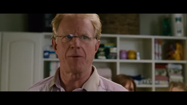 A middle-aged man with blond hair and wire-rimmed glasses gives a deadpan, skeptical expression in what appears to be a movie or TV scene set in a room with shelving in the background.