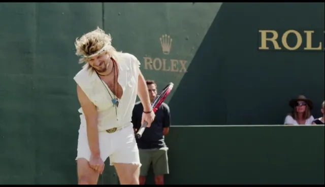 A man dressed in a retro 1970s-style tennis outfit with a headband, long blonde hair, and a necklace holds a racket on a tennis court with Rolex branding visible in the background. He appears to be reacting to a missed shot or bad play with a dejected posture.