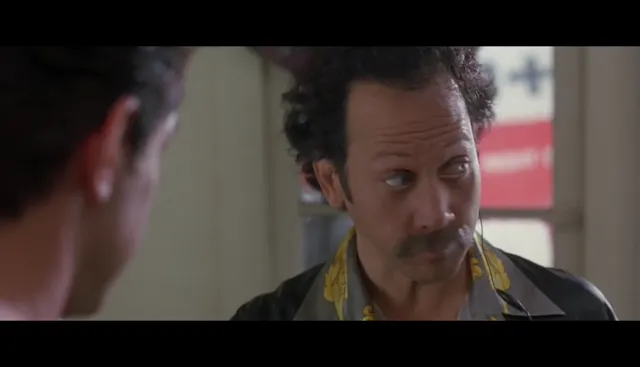 A curly-haired man with a mustache wearing a jacket with yellow trim stares intently at another person whose back is to the camera, in what appears to be a tense confrontational scene from a film.