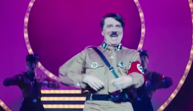 A person in a Hitler costume laughs and dances on a colorful stage with backup dancers, resembling a satirical musical performance. The theatrical setting with bright lights and a circular backdrop suggests a parody or comedy show context.
