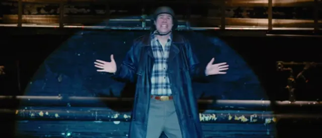 A man wearing a dark leather coat, plaid shirt, and hat stands with arms spread wide in an expressive, theatrical pose in front of what appears to be a large vehicle or structure at night. The scene has a cinematic, movie-still quality suggesting a dramatic or comedic moment.