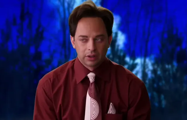 A man wearing a dark red button-up shirt with a matching striped tie and pocket square looks downward with a dejected or disappointed expression, set against a blue forest backdrop. The scene appears to be from a TV show or skit, possibly a news or interview segment parody.