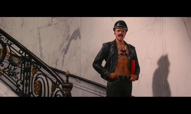 A mustachioed man wearing an open leather jacket with a harness, leather cap, and holding a red object stands confidently at the base of an ornate grand staircase with gilded ironwork. The scene appears to be from a 1970s or 1980s film with a campy or comedic tone.