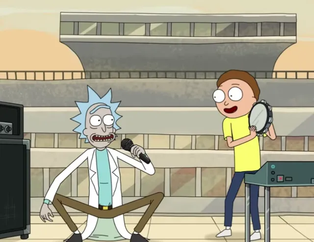 Rick sits cross-legged on a stage holding a microphone while Morty stands beside him holding a tambourine, both appearing to perform in an outdoor amphitheater setting. The scene is from the animated show Rick and Morty, capturing a lighthearted musical moment.