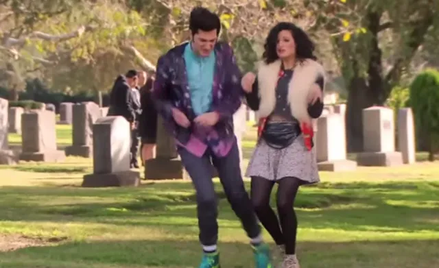 A young man and woman dance energetically together in a cemetery surrounded by gravestones, both dressed in colorful, eclectic outfits. The lighthearted dancing contrasts with the somber graveyard setting, suggesting a comedic or quirky TV show scene.