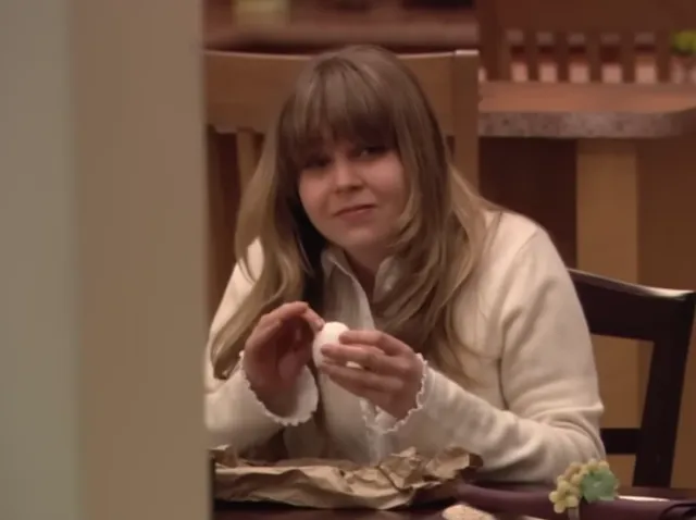 A young woman with long blonde hair and bangs sits at a dining table, smiling subtly while holding what appears to be a small food item, with grapes visible nearby. The scene appears to be from a TV show or film, captured in a warm indoor setting.