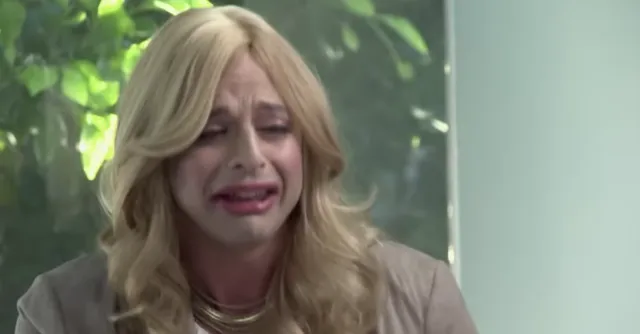 A blonde woman with long wavy hair is crying dramatically with a distressed facial expression, appearing to be in an emotional scene from a TV show or film. The setting looks like an indoor space with greenery visible through a window behind her.