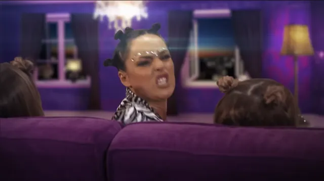 A woman with space buns and heavy makeup snarls aggressively over the back of a purple sofa, with two other people seated nearby. The purple-lit room and dramatic expression suggest a reality TV confrontation scene.