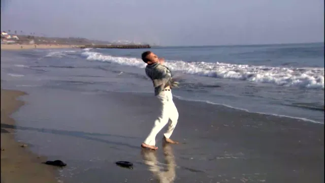 A man in white pants and a dark top dances expressively at the water's edge on a sandy beach, with ocean waves rolling in behind him. The scene has a carefree, joyful energy reminiscent of a TV show or movie clip.
