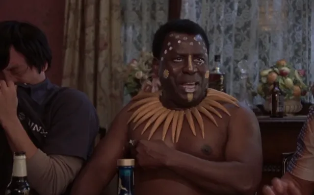 A shirtless man wearing tribal face paint and a large spiked neck collar sits indoors at what appears to be a party or gathering, with another person visible beside him looking away. The scene appears to be from a movie or TV show.