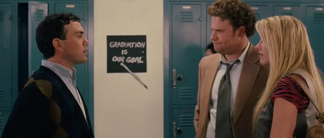 Two men and a woman face each other in a school hallway lined with blue lockers, with a chalkboard sign reading 'Graduation Is Our Goal' visible in the background. The scene appears to be from a comedy film, with a tense or confrontational interaction between the characters.