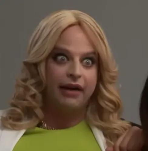 A man in drag wearing a blonde wig, green top, and white blazer makes an exaggerated wide-eyed shocked expression, likely from a comedy sketch or TV show. The face conveys extreme surprise or disbelief.