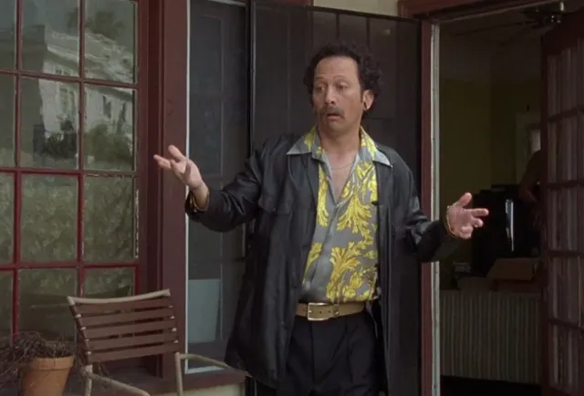 A man in a flashy yellow floral shirt and black leather jacket stands in a doorway with both hands raised in an exasperated or questioning shrug. The scene appears to be from a movie or TV show, capturing a moment of confusion or disbelief.