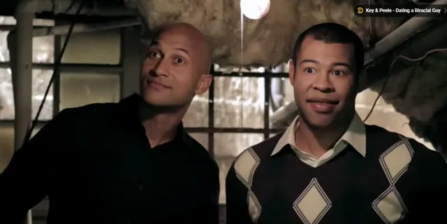 Keegan-Michael Key and Jordan Peele stand together in a dimly lit basement-like setting, both smiling at the camera in a scene from their Comedy Central sketch about dating a biracial guy.
