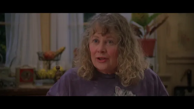 A middle-aged woman with curly blonde-gray hair wearing a purple shirt speaks in what appears to be a home kitchen setting. The scene looks like a still from a movie or TV show.