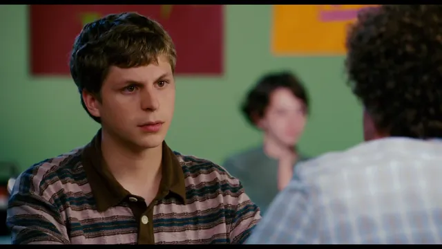 A young man in a striped shirt looks intently and somewhat skeptically at someone across from him, with another person visible in the background. The scene appears to be from a movie, likely Scott Pilgrim vs. the World, set in a casual indoor environment.