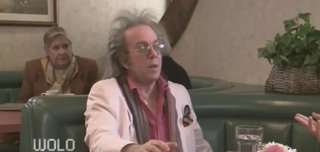 A wild-haired, eccentrically dressed man wearing glasses, a white blazer, and a colorful scarf gestures while speaking, apparently during a TV interview or news segment in a restaurant or lounge setting. An older woman sits in the background, and a 'WOLO' station watermark is visible.
