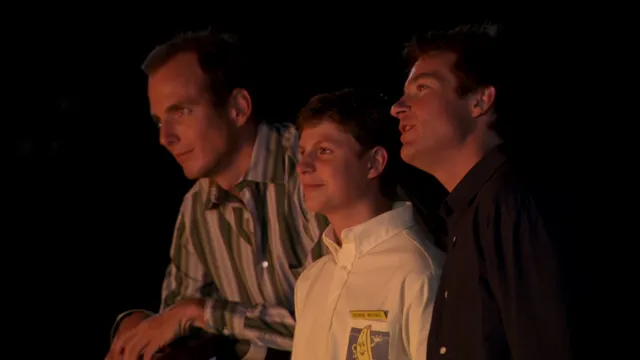 Two adult men and a young boy stand together in a dark setting, all gazing upward with expressions of wonder or awe, lit by warm light from below. The scene has a cinematic, nostalgic feel reminiscent of a movie still.
