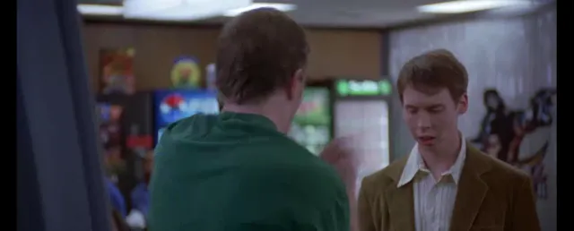 Two young men face each other in what appears to be an indoor public space, possibly a mall or arcade, with one wearing a green shirt and the other a tan blazer. The scene looks like a tense or serious conversation from a movie.