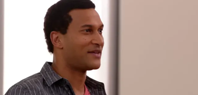A man in a striped shirt and pink undershirt smiles while looking to the side, appearing pleased or amused in what looks like a TV show or film scene.