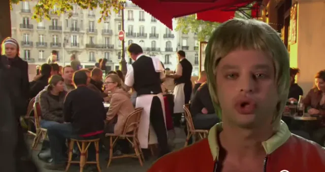 A man wearing a shaggy blonde wig and a red jacket stands in front of a busy Parisian outdoor café terrace, looking surprised or mid-speech. The scene appears to be a comedic TV show or skit set in Paris.