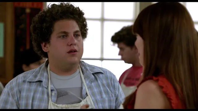 A young curly-haired man wearing an apron over a plaid shirt looks attentively at a woman with auburn hair in what appears to be a classroom or cooking setting. The scene appears to be from the movie Superbad.
