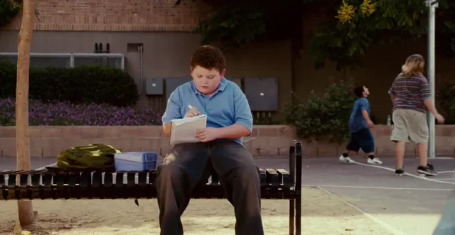 A young boy in a blue polo shirt sits alone on a bench at a school playground, writing intently in a notebook while other kids play in the background. The scene conveys loneliness or focused determination during recess.