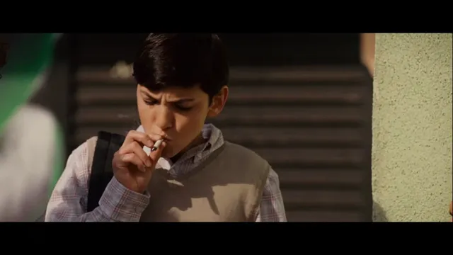 A young boy in a school vest and collared shirt is smoking a cigarette outdoors, with a serious or troubled expression on his face. The scene appears to be a still from a film or TV show.